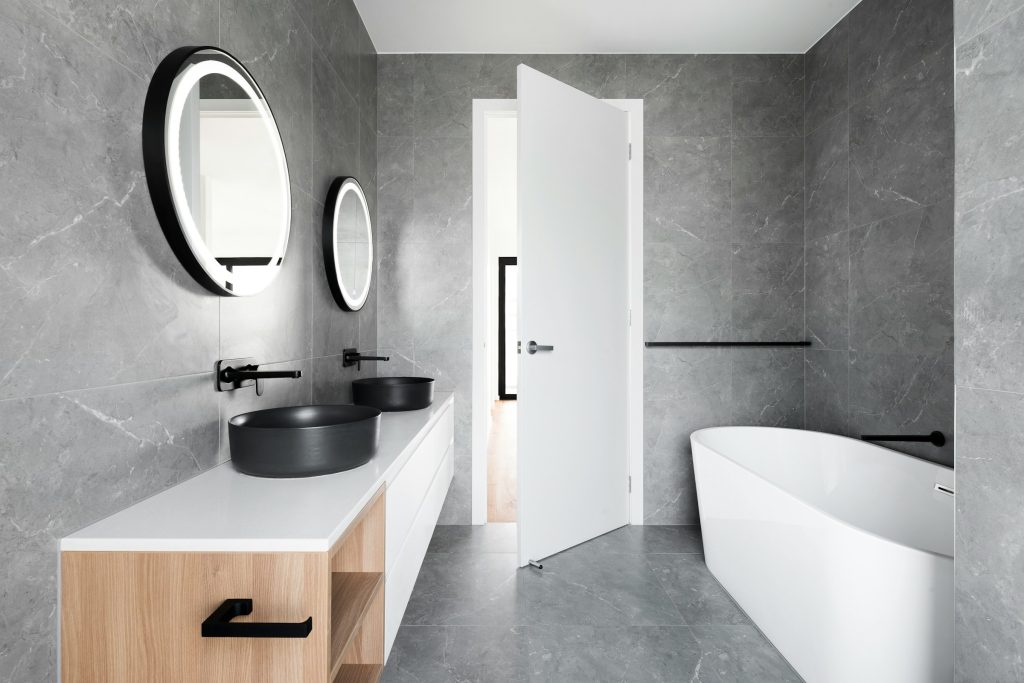 Grey 24x48 tiles with natural finish installed on a wall of bathroom which has a small wood vanity and a bathtub.